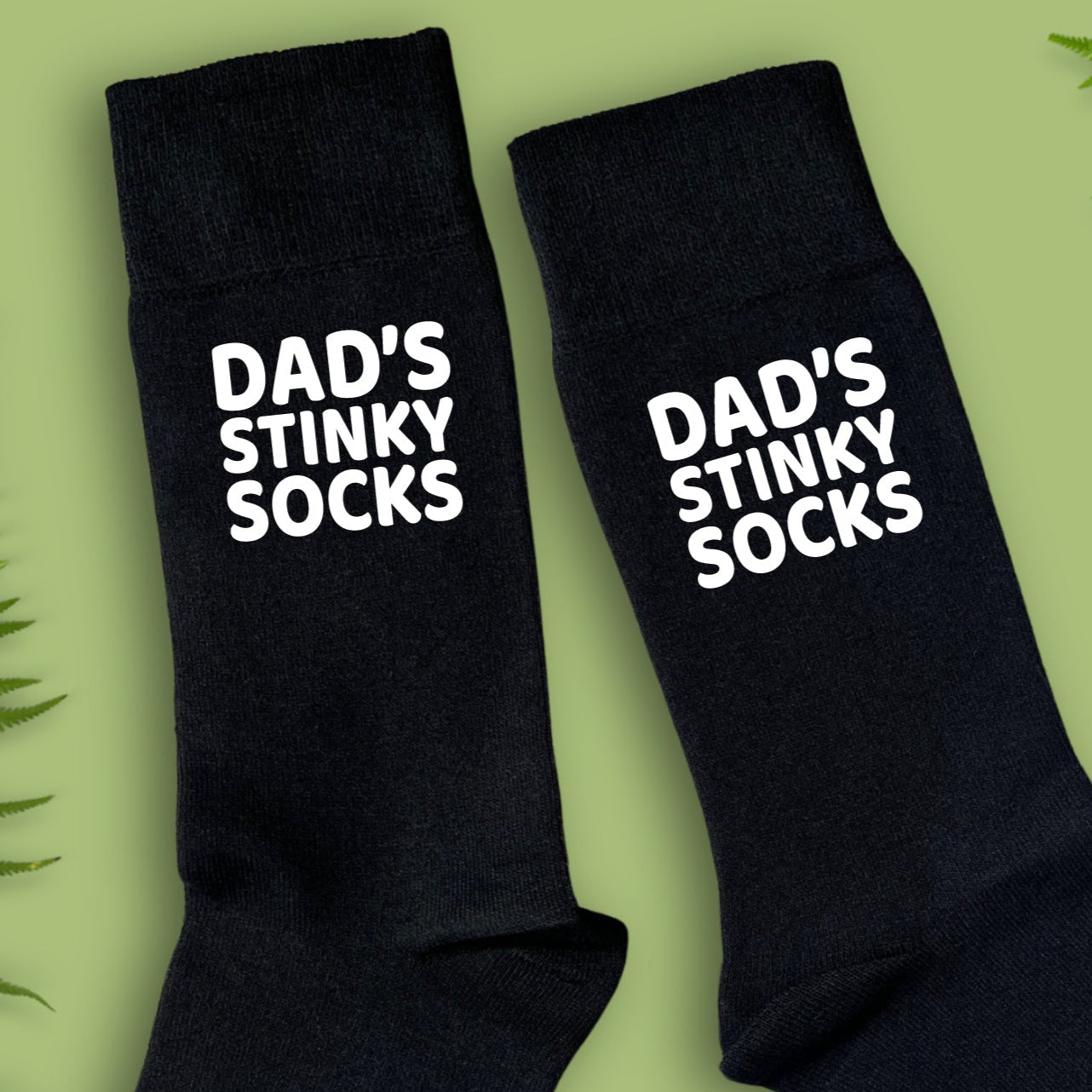 Close up of black socks with 'DAD'S STINKY SOCKS' text on a green background with fern leaves.