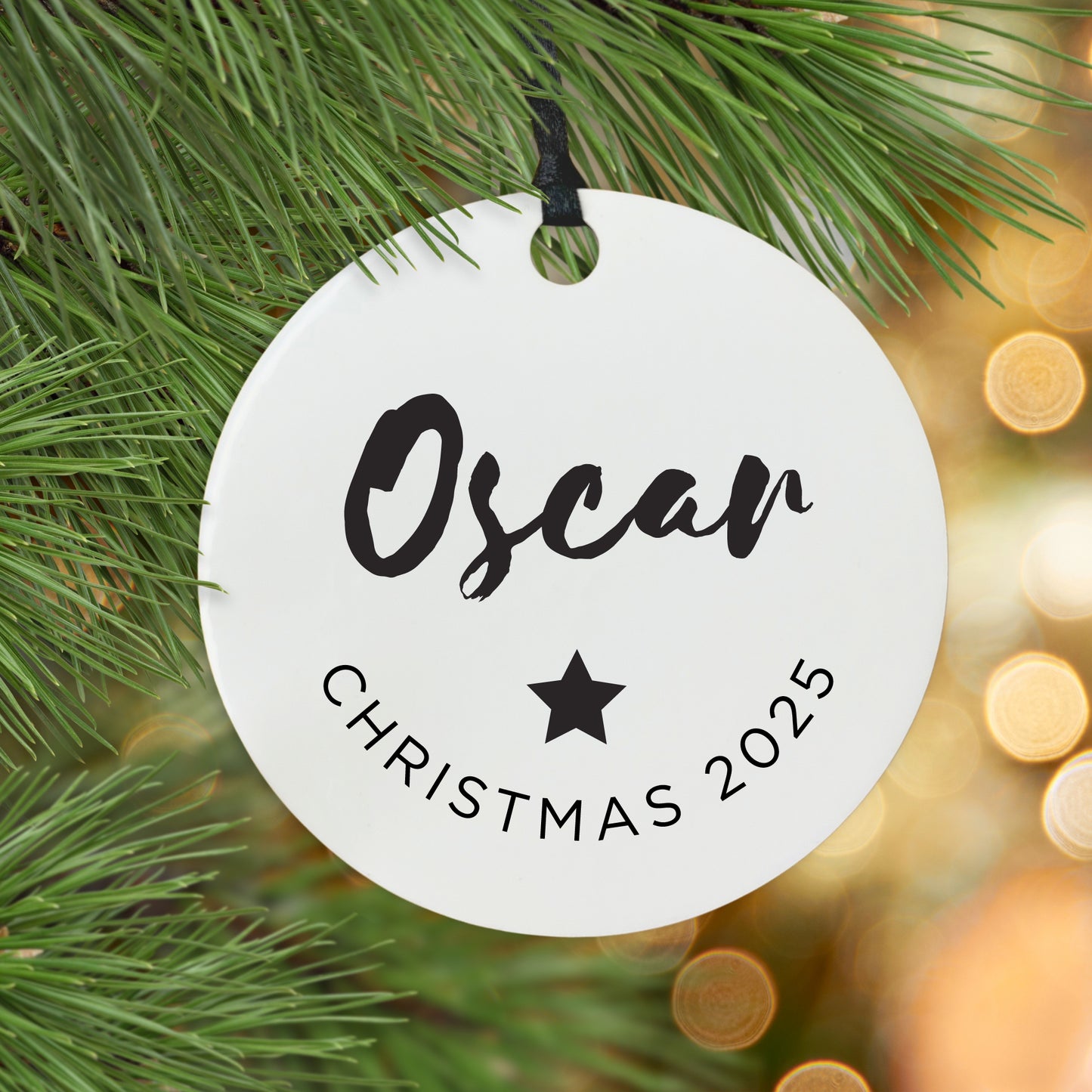 Round white ornament with name and Christmas 2025 text, hanging on a Christmas tree.