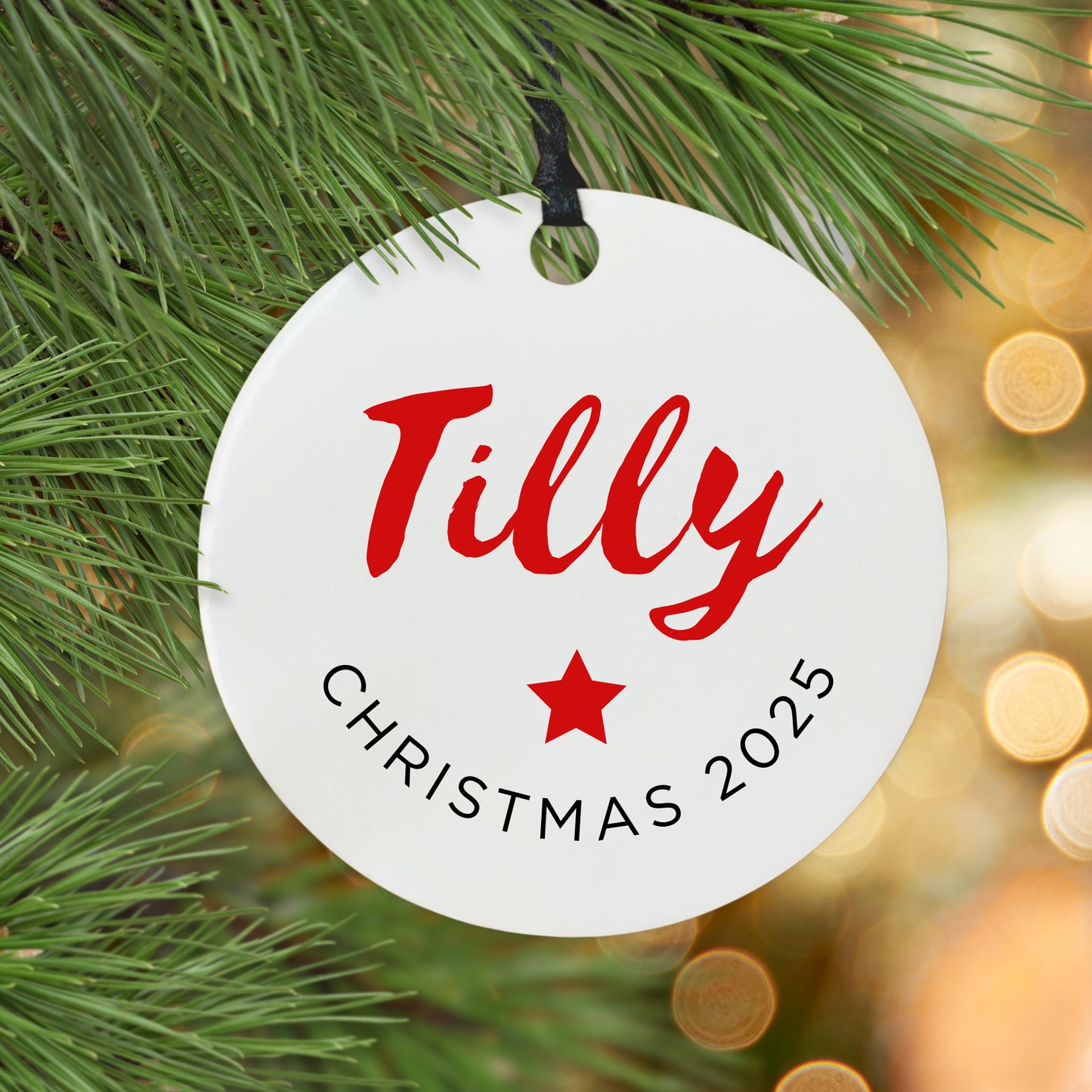 Round white ornament with red text name and 'Christmas 2025' on a Christmas tree branch.